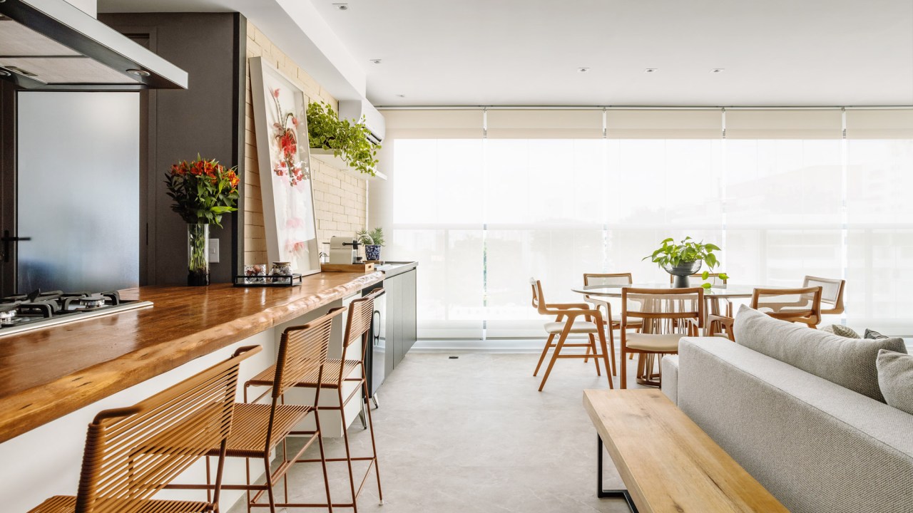 Sala integrada com varanda e cozinha; piso vinílico marmorizado, mesa redonda, bancada de madeira e sofá.