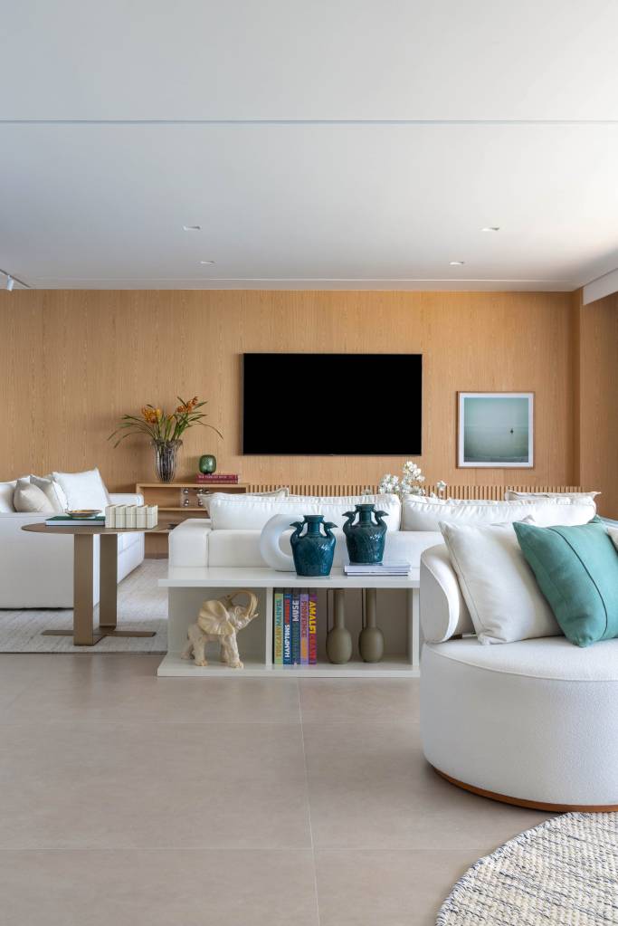 reforma-ape-365m-integra-estar-jantar-cria-copa-330arquitetura-02-sala-porcelanato Sala de estar clara com piso de porcelanato, sofás brancos, tv e paredes revestidas de madeira.