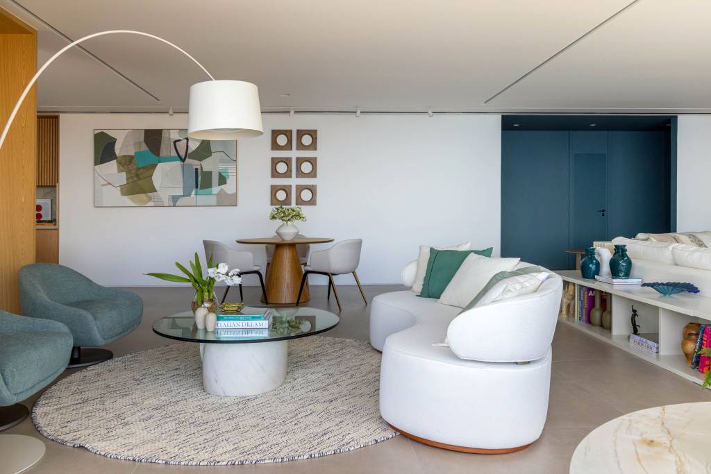 reforma-ape-365m-integra-estar-jantar-cria-copa-330arquitetura-13-sala Sala de estar com piso de porcelanato, sofá curvo branco, luminária de piso e tapete felpudo redondo.