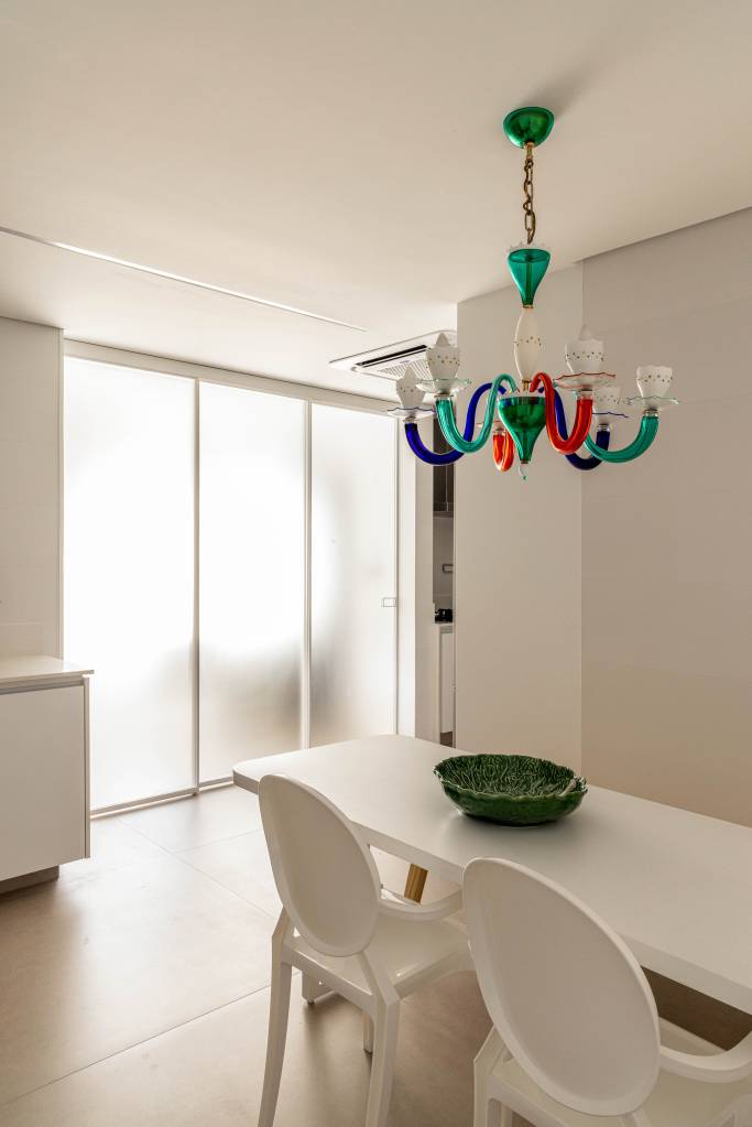 reforma-ape-365m-integra-estar-jantar-cria-copa-330arquitetura-18-jantar-branco Sala branca com mesa e cadeiras brancas e lustre de vidro colorido.