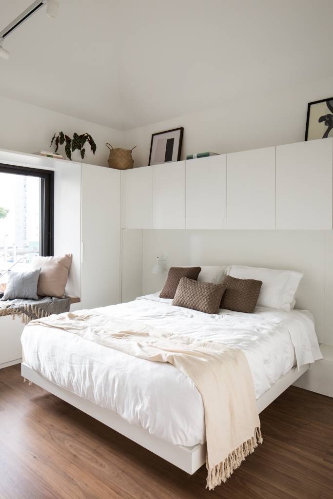 Quarto branco com armário branco em cima da cama e mesa lateral.