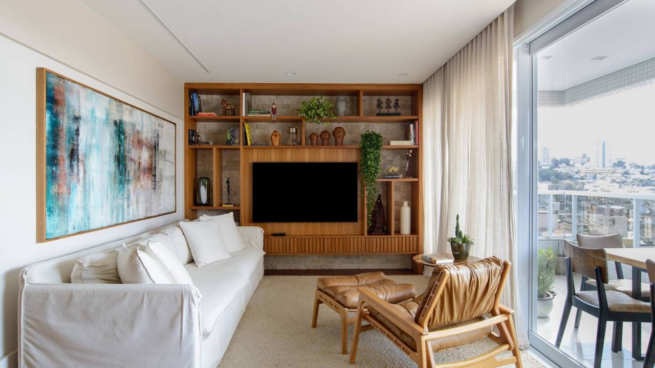 Apê de 140 m² feito para receber une madeira e mármore no décor. Projeto de Letícia Nannetti. Na foto, sala de estar com varanda, estante e parede de pedra.