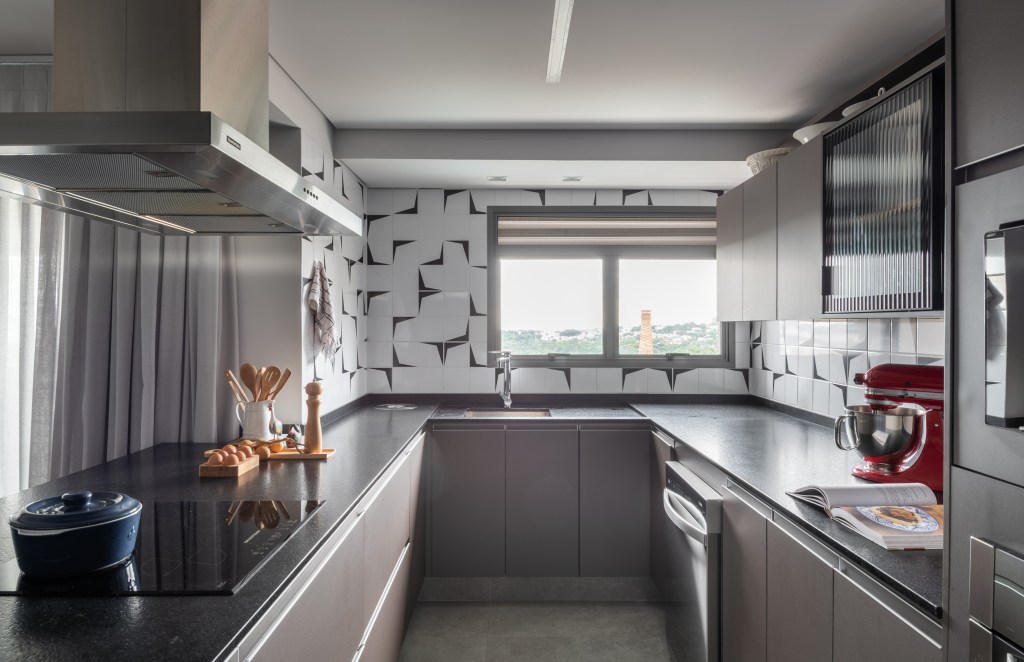Cozinha em formato de u com janela, bancada preta, armários taupe e parede de azulejos.