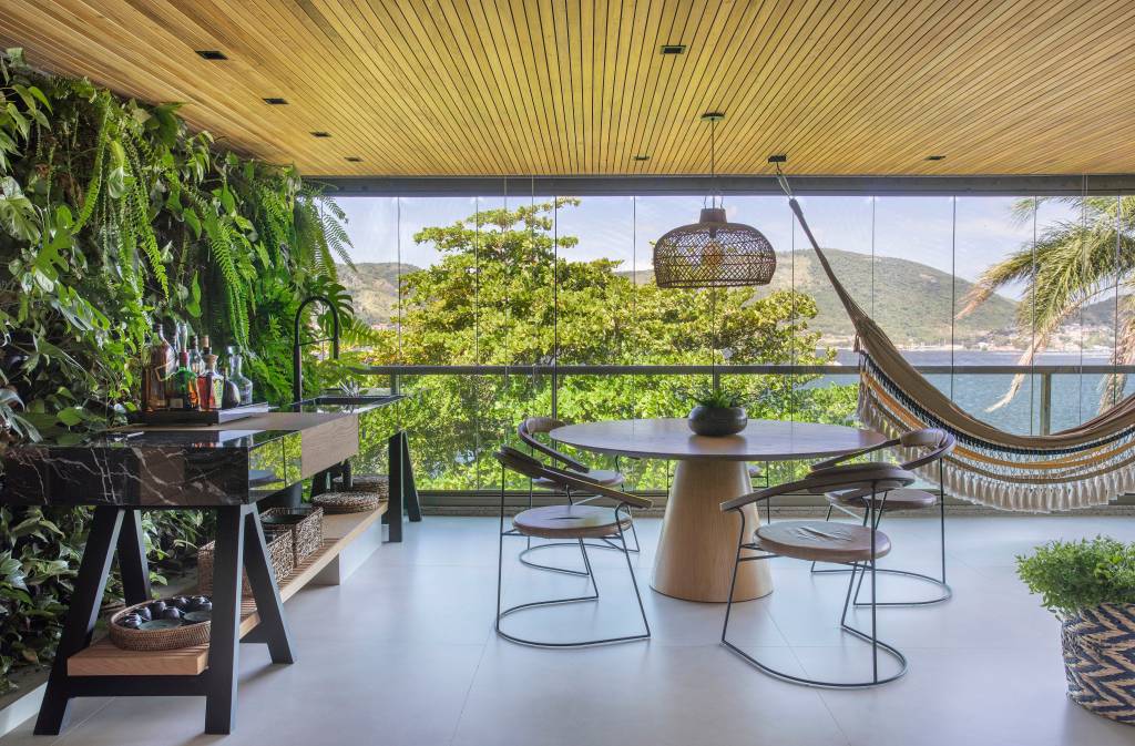 ape-220m-area-gourmet-rede-sala-quarto-varanda-integrada-03-jardim-vertical Varanda fechada com vidro, teto com ripas de madeira, mesa redonda de com cadeiras.