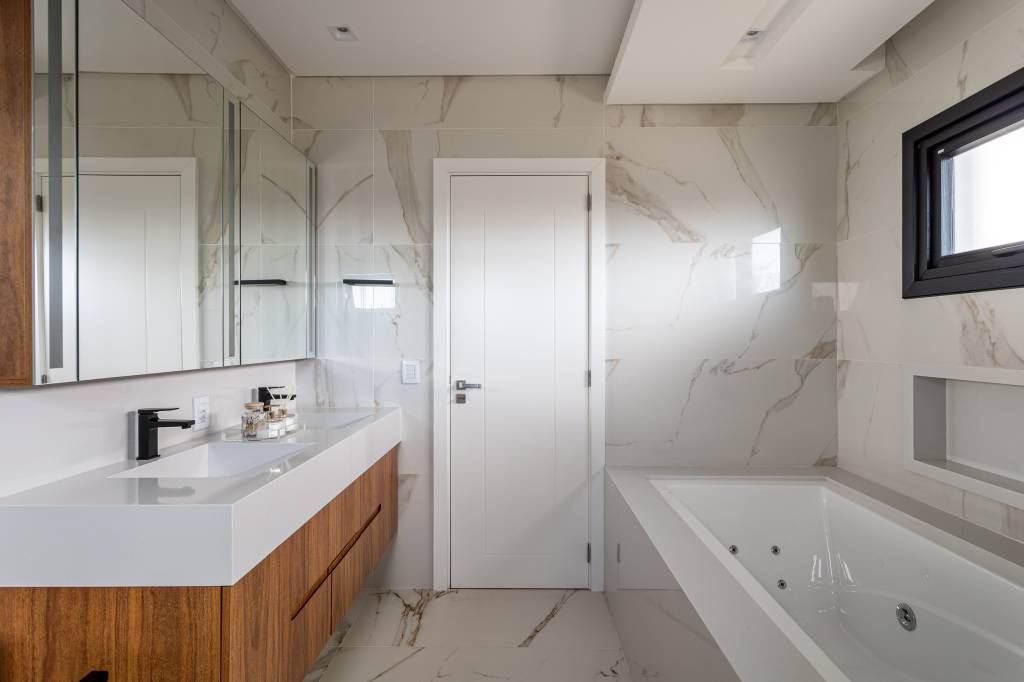 casa-246m-ilha-quartzo-cozinha-integrada-clare-arquitetura-12-banheiro-banheira Banheiro com duas cubas esculpidas brancas, armário em madeira, banheira e revestimento marmorizado.