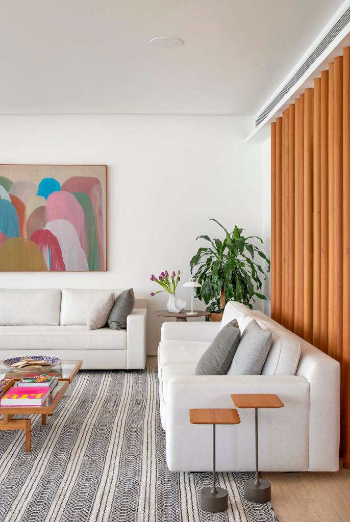 paleta-cores-claras-ripados-madeira-ape-275m-studio021arquitetura-04-sala Sala de estar branca com sofá branco, tapete listrado azul, banco branco e mesa de centro de madeira.