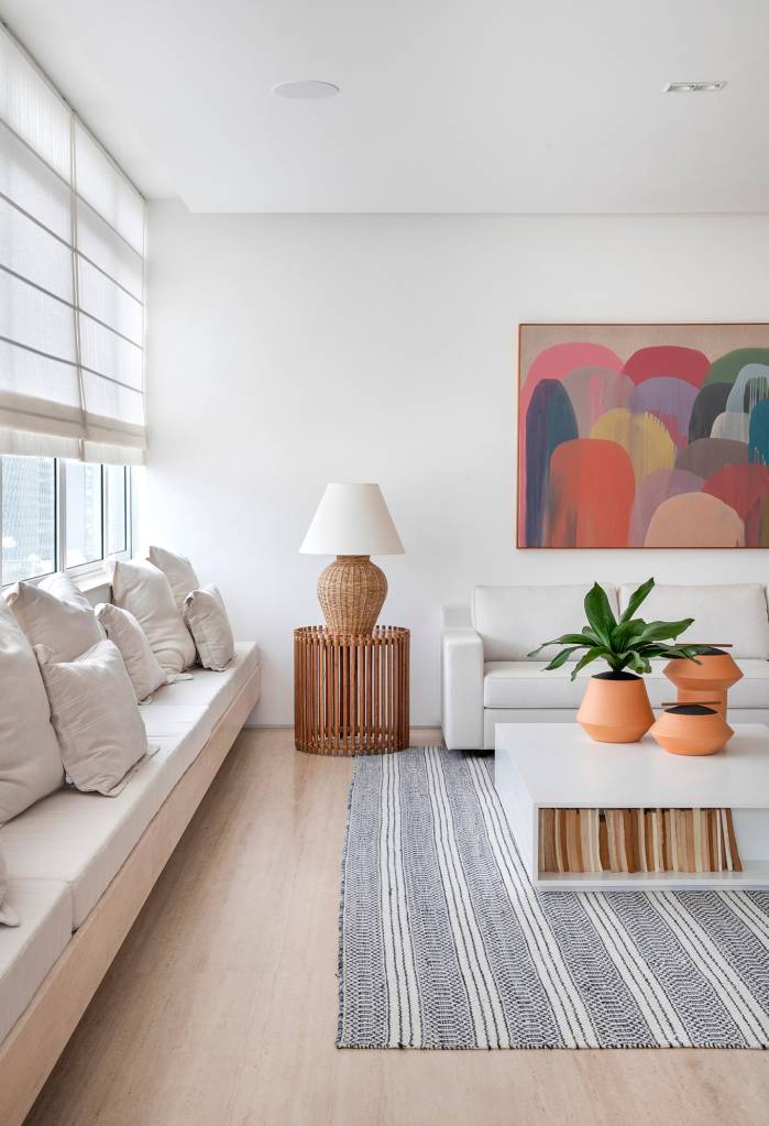 paleta-cores-claras-ripados-madeira-ape-275m-studio021arquitetura-07-sala-banco Sala de estar branca com sofá branco, tapete listrado azul, banco branco e mesa de centro de madeira.