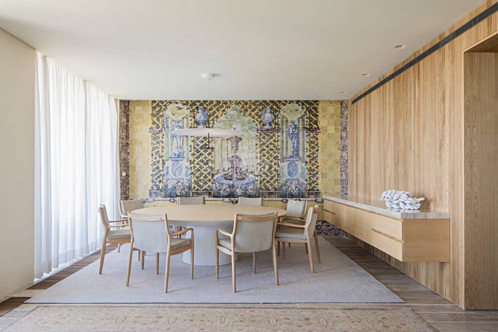 apartamento-portugal-tv-escondida-atras-quadro-deslizante-mauricio-nobrega-01-sala-jantar Sala de jantar com mesa de madeira redonda, painel de azulejos antigos ao fundo e tapetes.