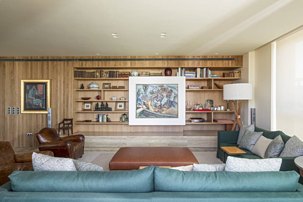 apartamento-portugal-tv-escondida-atras-quadro-deslizante-mauricio-nobrega-10 Sala de estar com estante de madeira com quadro que desliza, sofá verde, poltronas vintage de couro e tapetes.