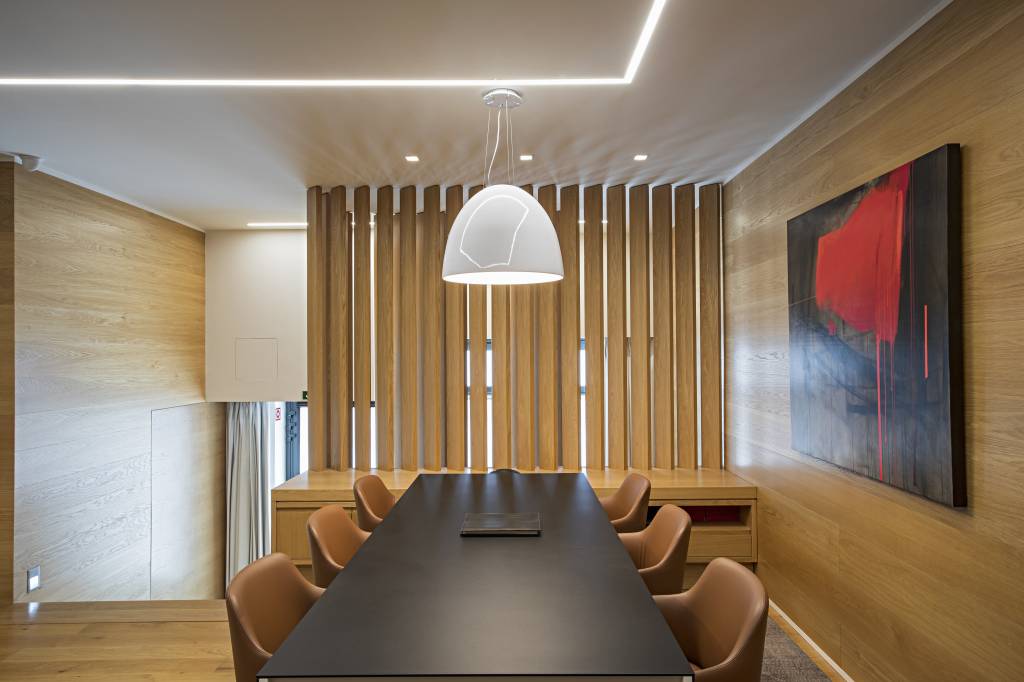 apartamento-portugal-tv-escondida-atras-quadro-deslizante-mauricio-nobrega-19 Escritório com paredes revestidas de madeira, mesa preta, brises de madeira, luminária e sanca iluminada.