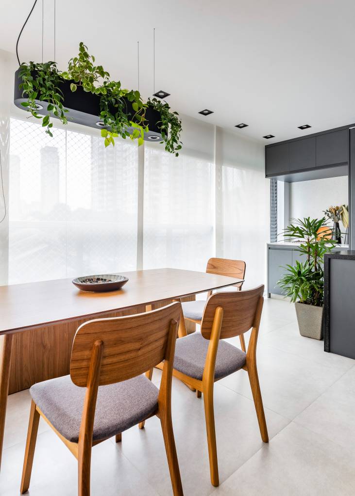 Sala de jantar em varanda integrada fechada com vidro com mesa de madeira, banco e luminária com plantas suspensas.