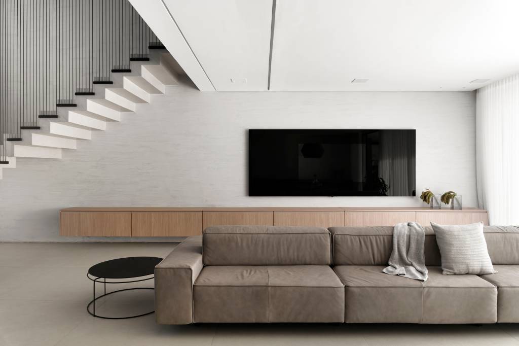 minimalista-casa-665m-porcelanato-paleta-preta-branca-piacesiarquitetura-01 Sala de tv minimalista com parede cinza clara, rack de madeira e sofá baixo; escada branca com guarda corpo preto.