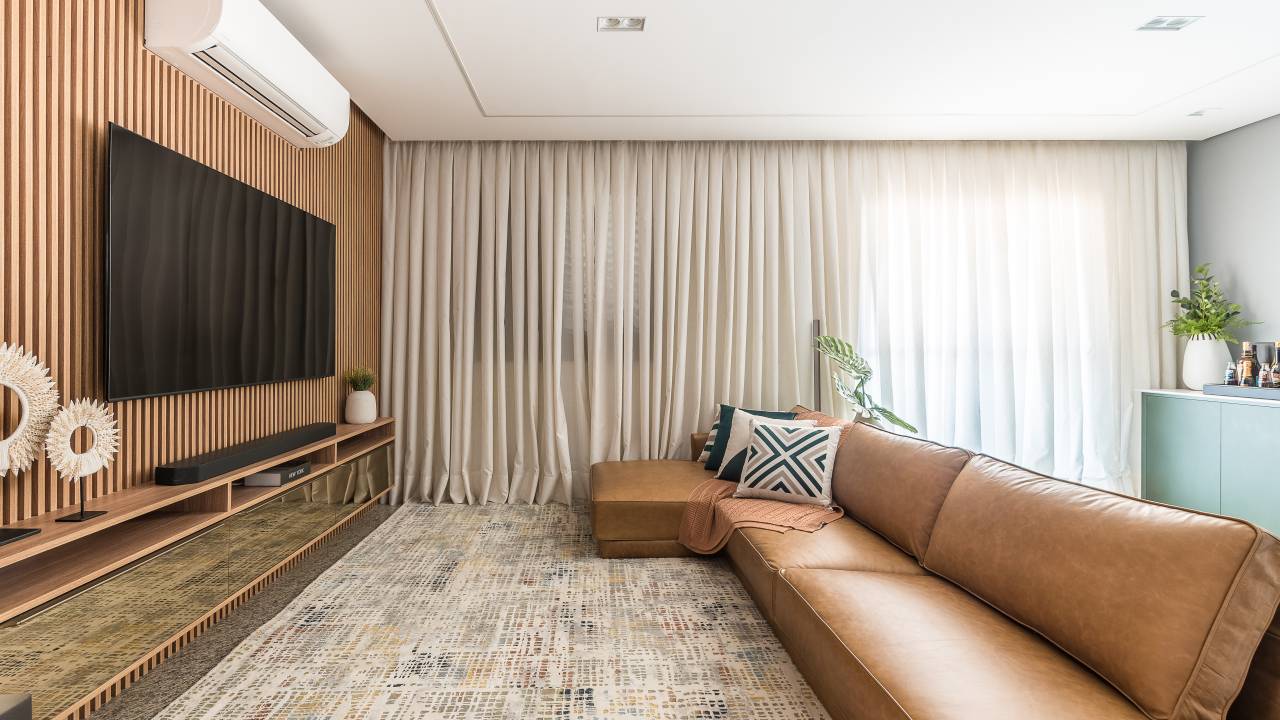 Projeto de Clareira Arquitetura. Na foto, sala de estar com painel ripado, sofá de couro e cortina.