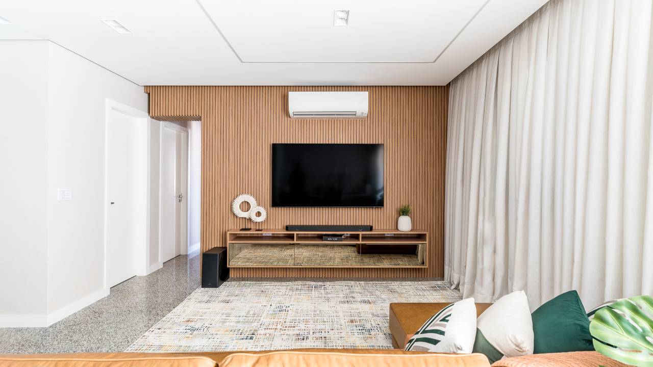 Projeto de Clareira Arquitetura. Na foto, sala de estar com painel ripado, sofá de couro e cortina.