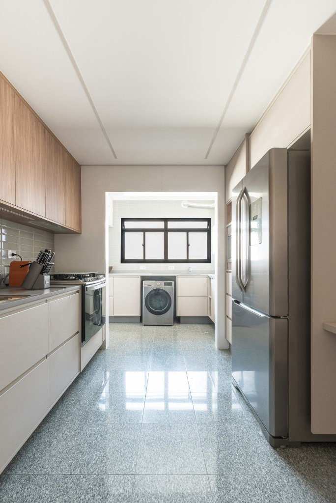 Projeto de Clareira Arquitetura. Na foto, cozinha com piso de granito, marcenaria bege e backsplash de azulejos. Lavanderia ao fundo.