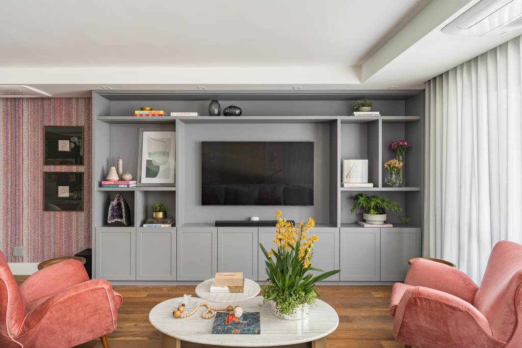 reforma-cria-suite-closet-integrado-spaco-interior-03-sala-tv-estante Sala de estar com piso de madeira, papel de parede e estante cinza com nichos. Sofá claro e poltronas laranja.