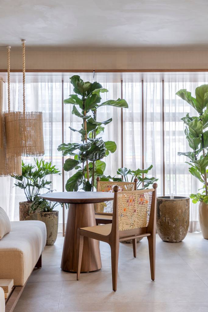 Sala de estar em tons beges, mesa redonda pequena de madeira, cadeiras de palhinha, luminária de palhinha e plantas.