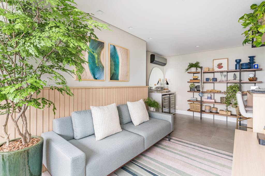 Apartamento de 60 m² ganha décor delicado repleto de madeira e plantas. Projeto de Bia Hajnal. Na foto, sala de estar com sofá cinza e lambri de madeira., Arvore em vaso. Estante com lembranças de viagem.