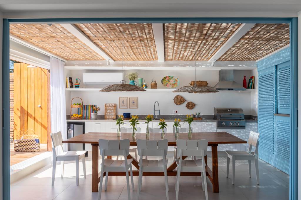 Projeto de Brise Arquitetura. Na foto, área gourmet com cobertura de pergolado com palhinha, mesa de madeira, cadeiras brancas e bancada de cozinha.