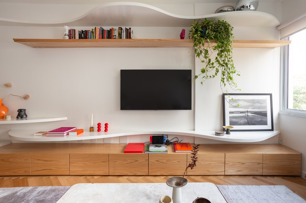 Concreto, verde e rosa marcam o décor deste apê de 110 m². Projeto de MNBR Arquitetos. Na foto, sala de TV com prateleira curva, plantas e livros.