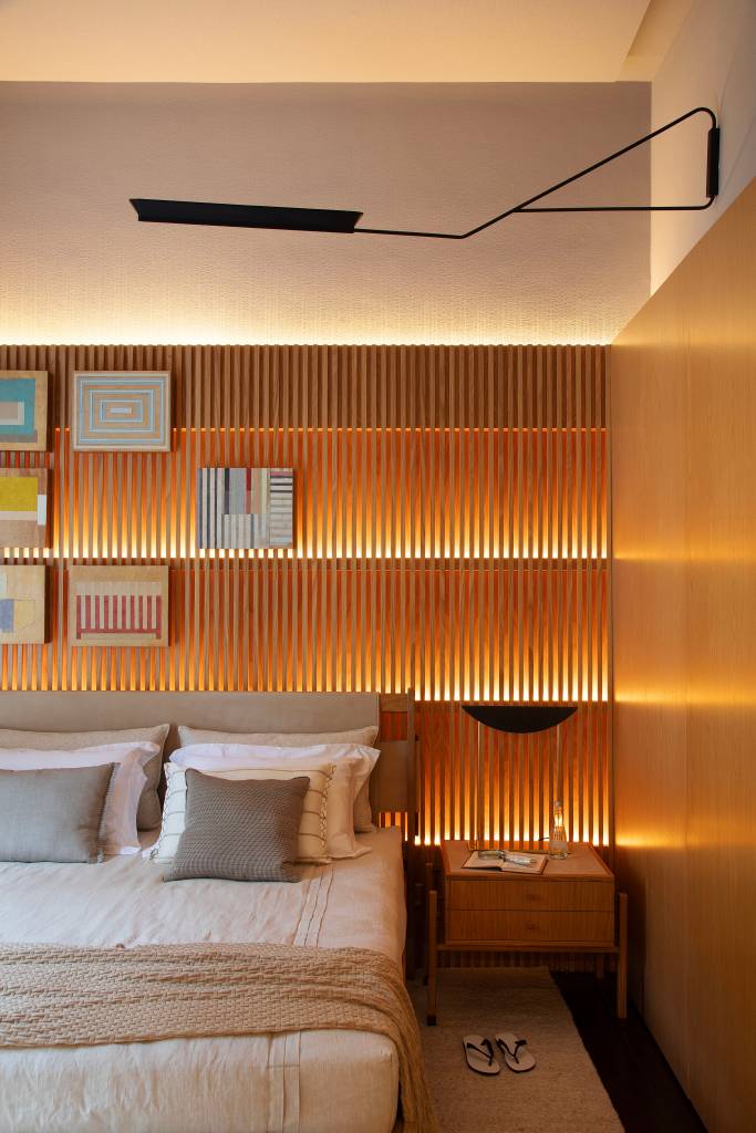 madeira-design-brasileiro-quarto-de-hotel-studio021arquitetura-casacor-rio-2023-10 Quarto 021 - Projeto do Studio021 Arquitetura para a CASACOR Rio 2023. Na foto, quarto com cama e parede ripada com quadros iluminada.