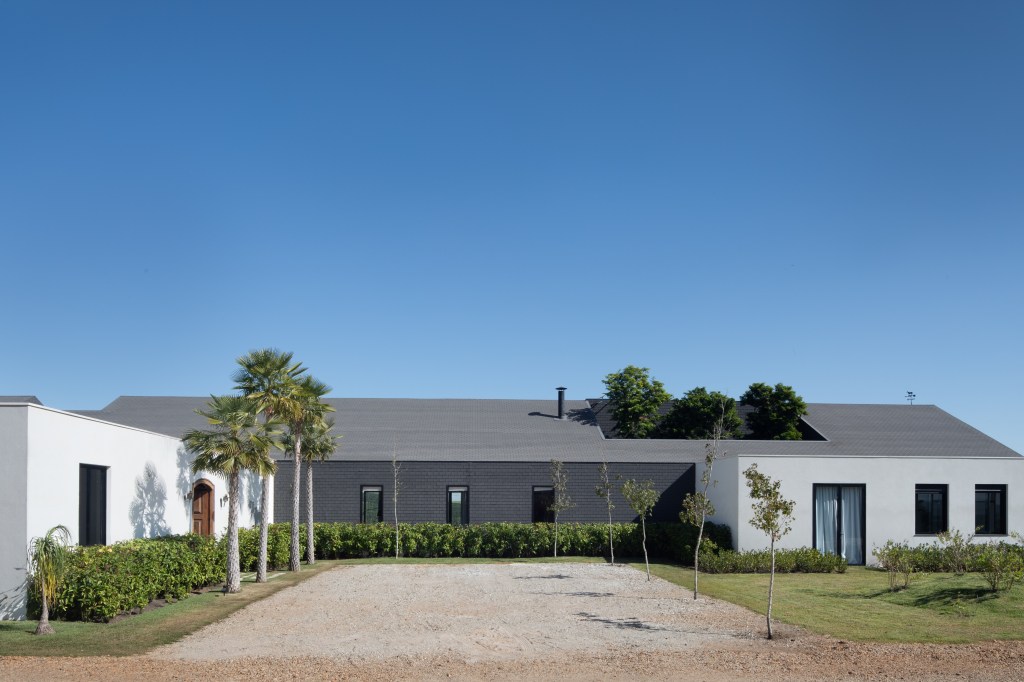 Casa celeiro de madeira une o rústico e o moderno em projeto de 1230 m². Projeto de Otto Felix. Na foto, fachada da casa com palmeiras e muro branco.