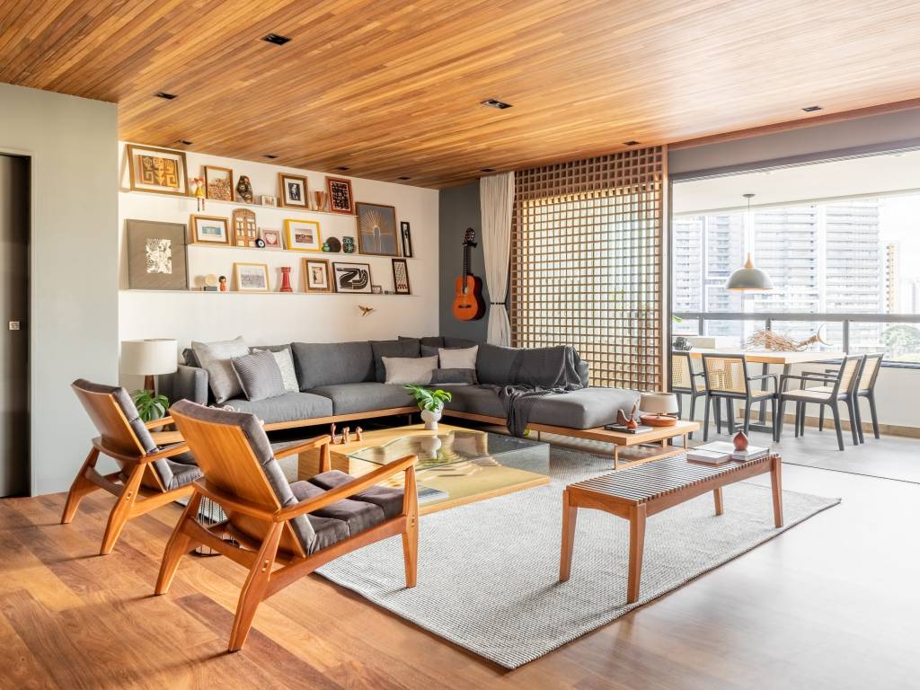 Cozinha integrada com bancada de neolith e cristaleira é destaque em apê. Projeto de Pietro Terlizzi. Na foto, sala com forro de madeira, prateleira com livros, sofá cinza e muxarabi dividindo varanda.