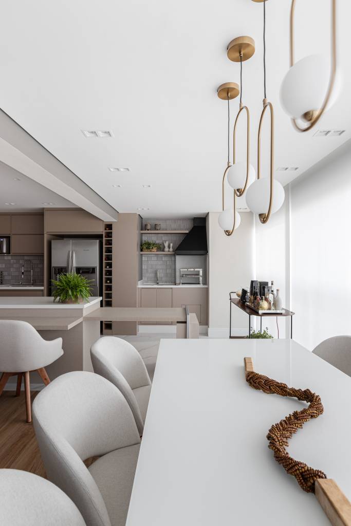 Reforma em apê de 140m² transforma banheiro em biblioteca de home office. Projeto de Blaia e Moura Arquitetos. Na foto, varanda gourmet integrada com mesa branca e cozinha com marcenaria bege.