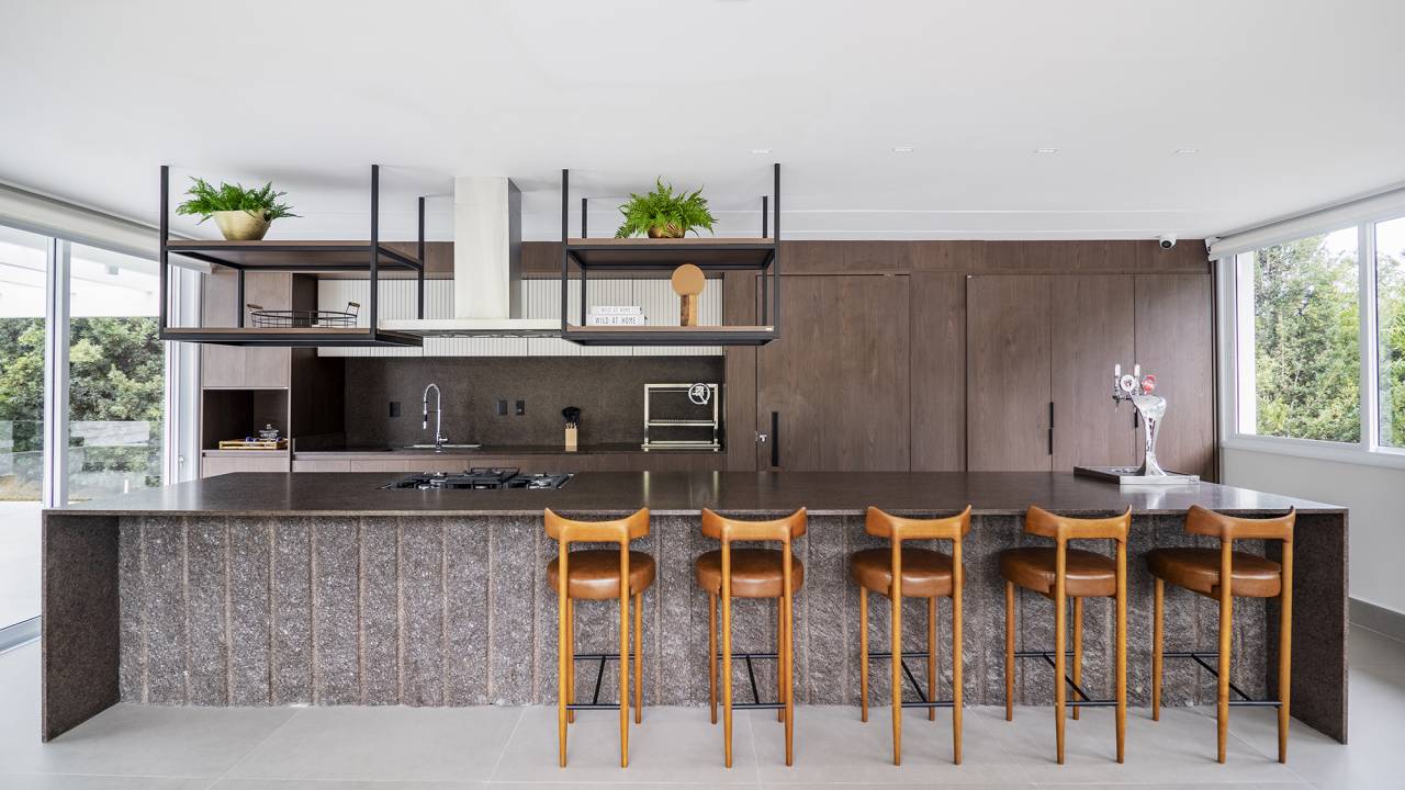 Área gourmet integrada ao jardim é o coração desta casa. Projeto de TM Arquitetura. Na foto, cozinha com ilha, marcenaria escura e banquetas.