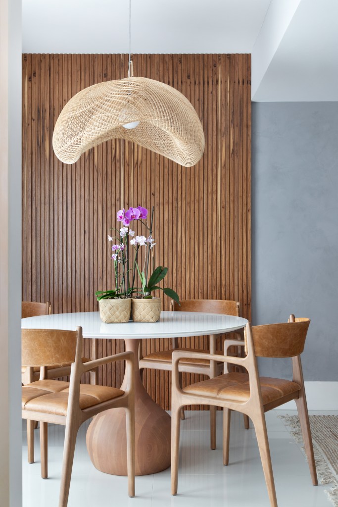 Couro, madeira e fibra compõem décor contemporâneo de cobertura de 130 m². Projeto de Travessa Arquitetura. Na foto, sala de jantar, parede ripada, mesa redonda branca, cadeira de madeira e couro, luminária de rattan.