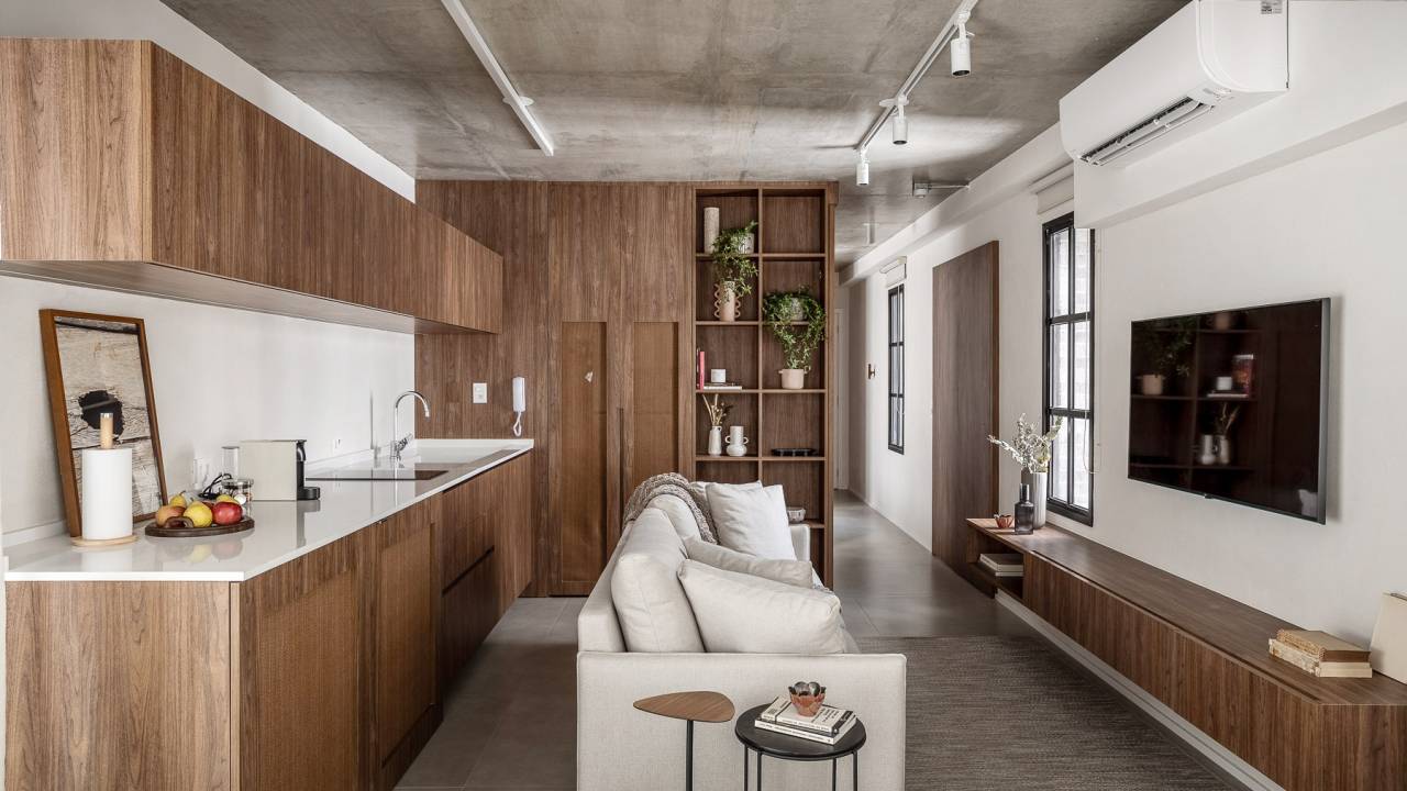 Marcenaria personalizada otimiza espaço e esconde eletros em apê de 48 m². Projeto de Carolina Gava. Na foto, sala integrada com cozinha, sofá branco, piso de porcelanato.