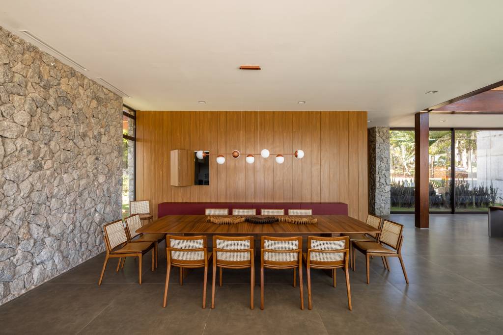 casa-composta-por-tres-volumes-ligados-por-pergolado-de-madeira-arkitito-13 Casa é composta por três volumes ligados por pergolado de madeira. Projeto de Arkitito. Na foto, sala de jantar, mesa de madeira, parede de pedra.