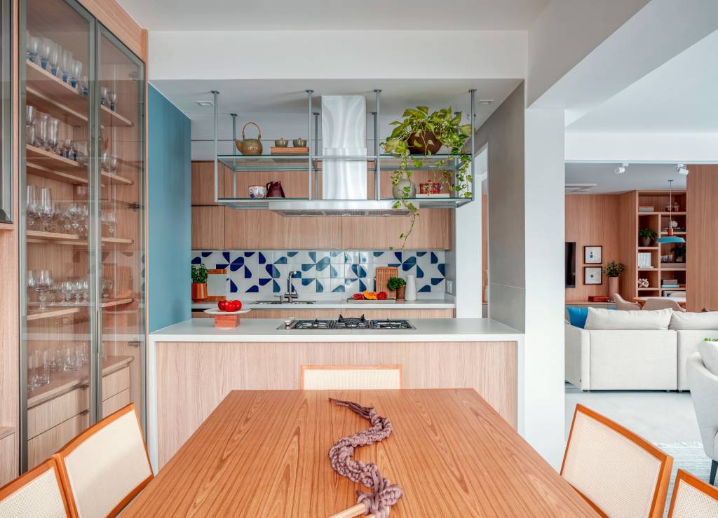 Cristaleira piso-teto rouba a cena na sala de jantar desta cobertura. Projeto de Paula Muller. Na foto, cozinha integrada, bancada branca, mesa de jantar de madeira retangular, backsplash de azulejos.