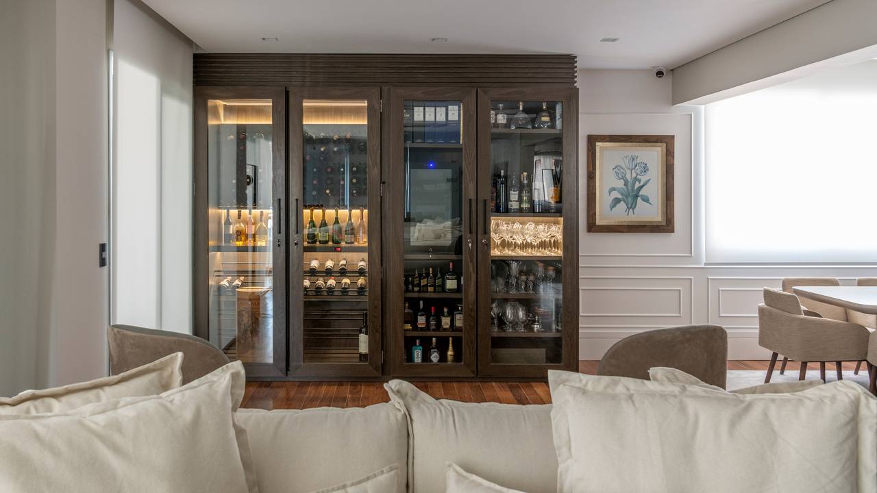 Madeira escura e vista para skyline trazem elegância para apê de 280 m². Projeto Studio Elã. Na foto, sala de estar com adega, sofás e paredes amadeiradas.