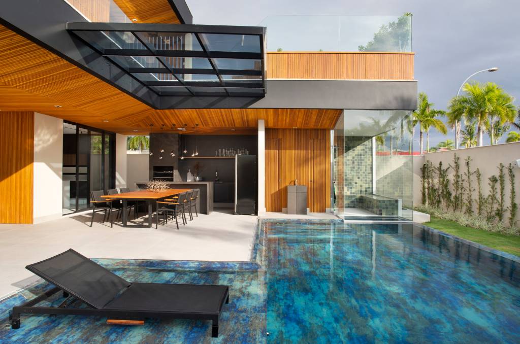 Painéis ripados e área gourmet convidam ao lazer e aconchego nesta casa. Projeto de Studio MH Arquitetura. Na foto, área externa com piscina, área gourmet, churrasqueira e espreguiçadeira.