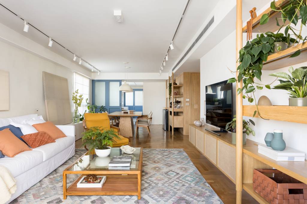Apto Brooklin Novo Apê de 180 m² ganha azul na marcenaria e nos revestimentos da cozinha. Projeto de Studio LAK. Na foto, sala de estar, tapete colorido, estante de madeira clara com plantas, sofá branco, mesa de centro.