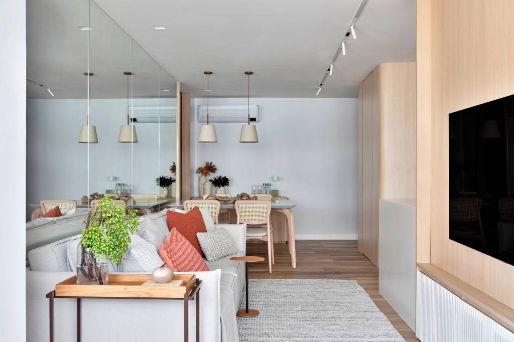 Espelho atrás do sofá amplia apê de 70 m² com paleta de cores clara. Projeto de FPR Studio. Na foto, sala de estar integrada com jantar, sofá cinza claro, cadeira com encosto de palhinha.