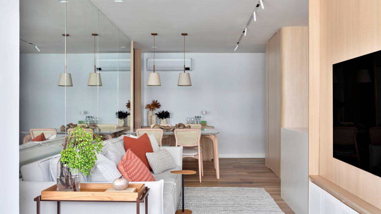 Espelho atrás do sofá amplia apê de 70 m² com paleta de cores clara. Projeto de FPR Studio. Na foto, sala de estar integrada com jantar, sofá cinza claro, cadeira com encosto de palhinha.