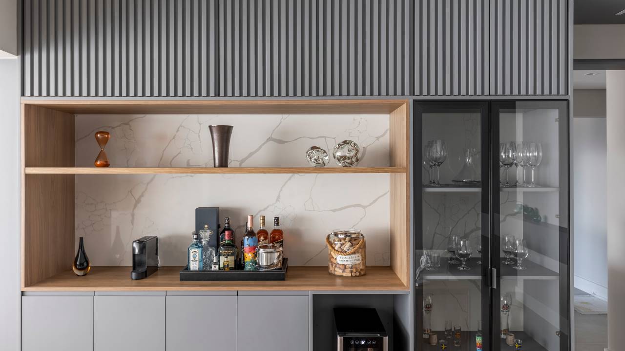 Varanda de apê tem dois espaços: um gourmet e outro de estar. Projeto de Blaia e Moura. Na foto, bar com marcenaria cinza, armário ripado, nicho e cristaleira.