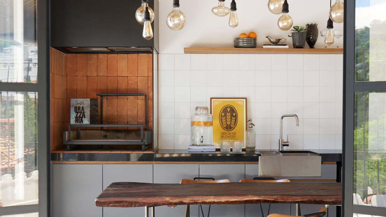 Porta de serralheria vira luminária pendente em cobertura com parrilheira. Projeto de Hugo Rapizo Arquitetura. Na foto, porta de vidro com moldura preta, piso de ladrilho preto e branco, área gourmet.