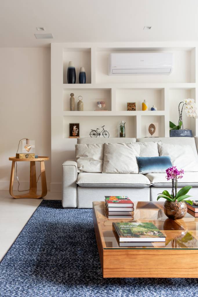 Reforma cria estante em gesso acartonado para acomodar tubulações. Projeto de Fernanda Medeiros. Na foto, sala, estante de nichos, ar condicionado, tapete azul, sofá branco.