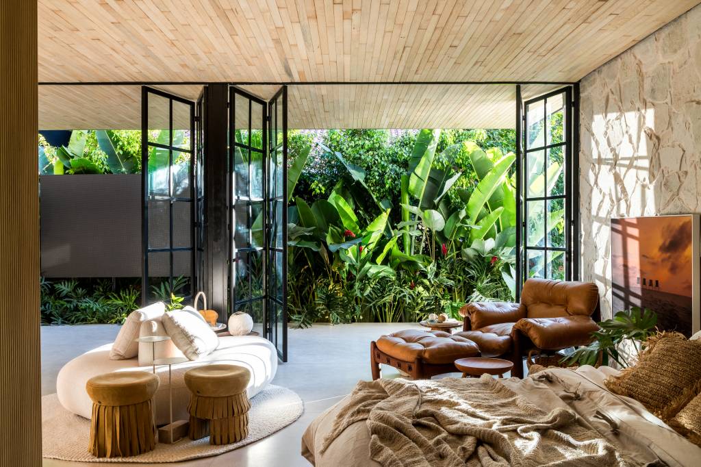 tiny-houses-discutem-sustentabilidade-relacao-com-natureza-casacor-pr-crippa-e-assis-arquitetura-05 3 tiny houses lindas que discutem sustentabilidade e a relação com a natureza. Projeto de Crippa e Assis para a CASACOR PR 2024. Na foto, sala, parede de pedra, poltrona com pufe, jardim ao fundo.