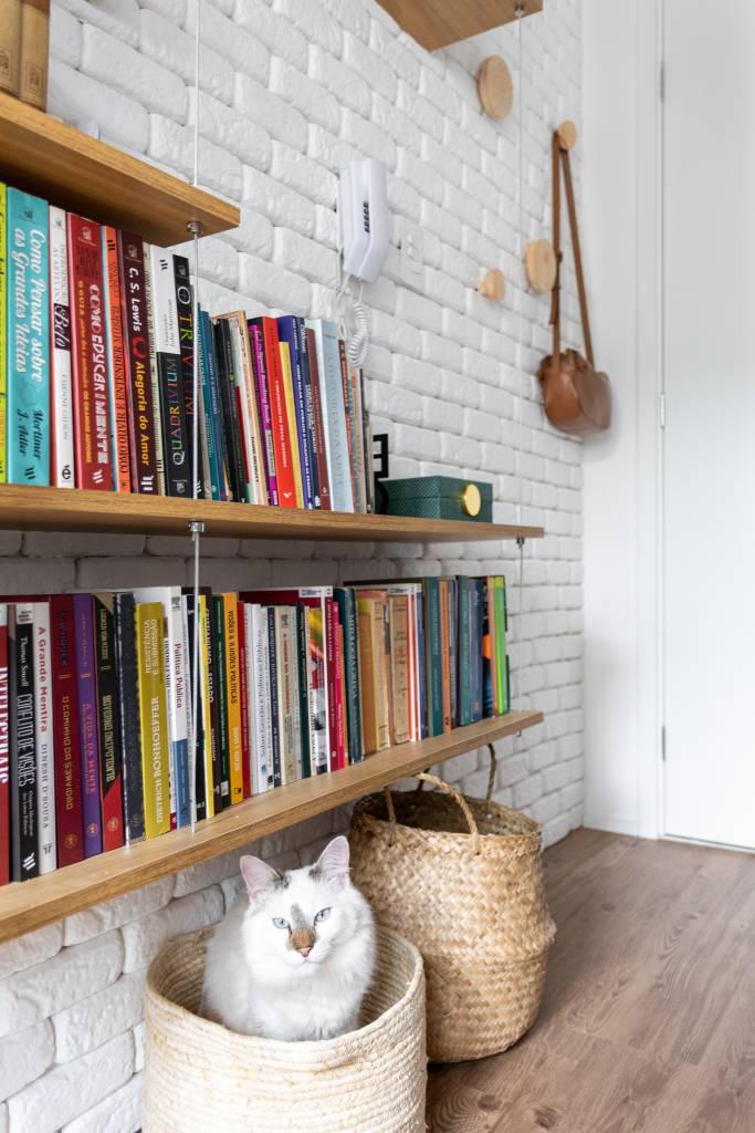 Miniapê de 25 m² tem sofá de canto alemão com baú e estante de mil livros. Projeto de Lemarchi Arquitetura. Na foto, prateleira, cesto, gato, parede de tijolinhos.