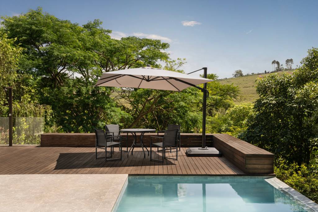 Casa de campo rústico contemporânea em Minas tem área gourmet voltada para piscina. Projeto de Camila Medrado. Na foto, piscina, ombrelone, vegetação.