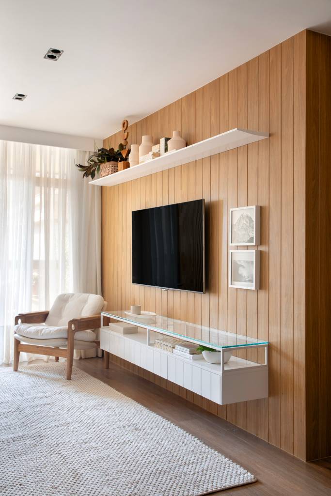 Tapete com trama artesanal, cortinas de linho e parede de pedras compõem apê de 120 m². Projeto de Ikeda Arquitetura. Na foto, sala de estar, tv, rack, painel de madeira, poltrona.