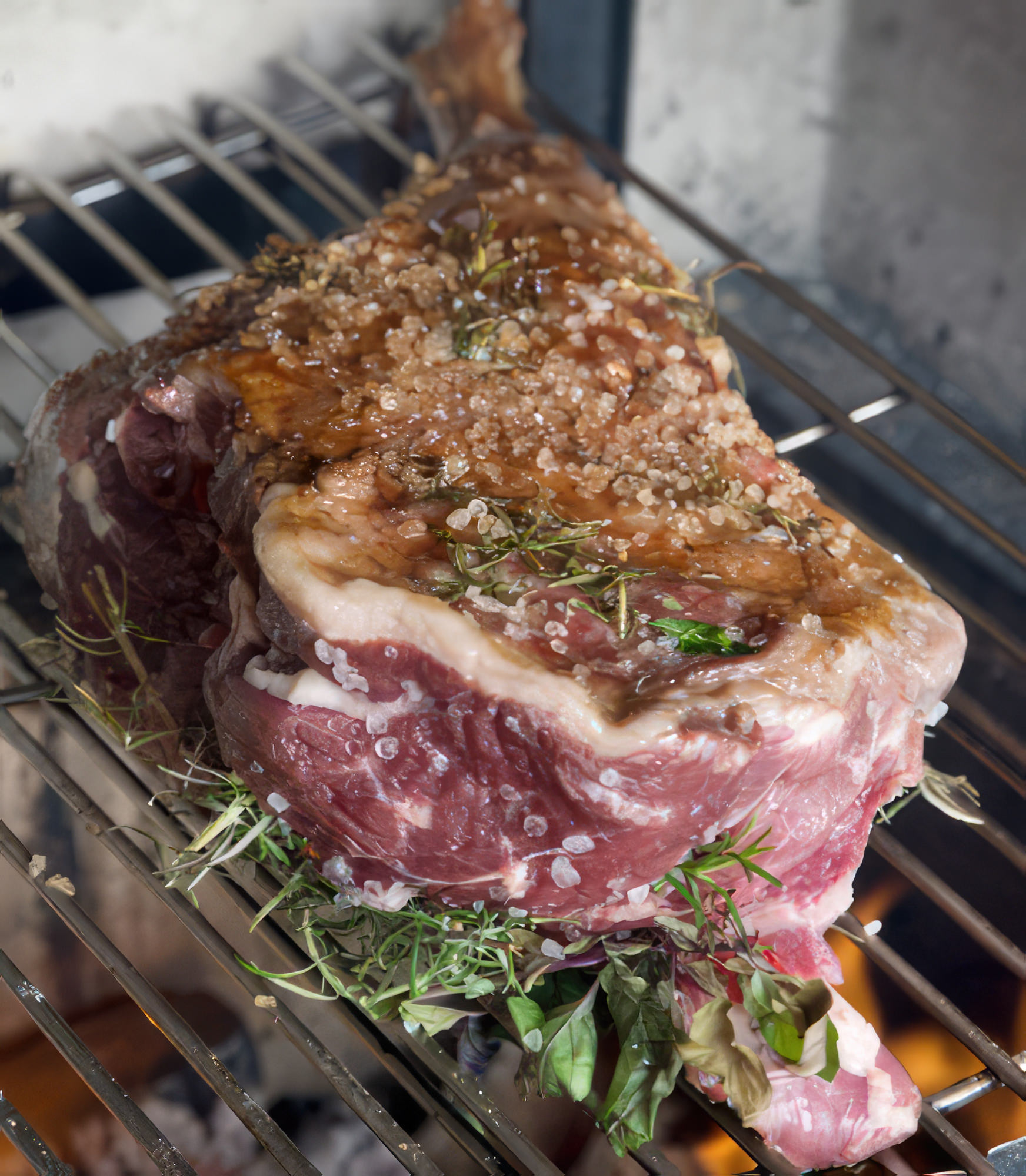 A imagem mostra um pernil de cordeiro sendo grelhado na churrasqueira, com uma crosta dourada e suculenta. O ambiente ao fundo indica uma reuni&atilde;o social, com pessoas apreciando o churrasco. Espetos e utens&iacute;lios de churrasco tamb&eacute;m s&atilde;o vis&iacute;veis, sugerindo um momento de prepara&ccedil;&atilde;o e confraterniza&ccedil;&atilde;o.