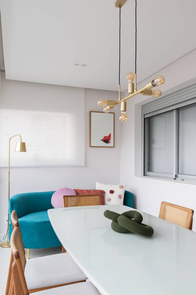 Móvel antigo funciona como bar e apoio para a mesa de jantar em apê de 69 m². Projeto de Studio Monfré. Na foto, varanda, mesa com tampo de vidro branco, luminária, móvel retrô, sofá curvo turquesa azul.