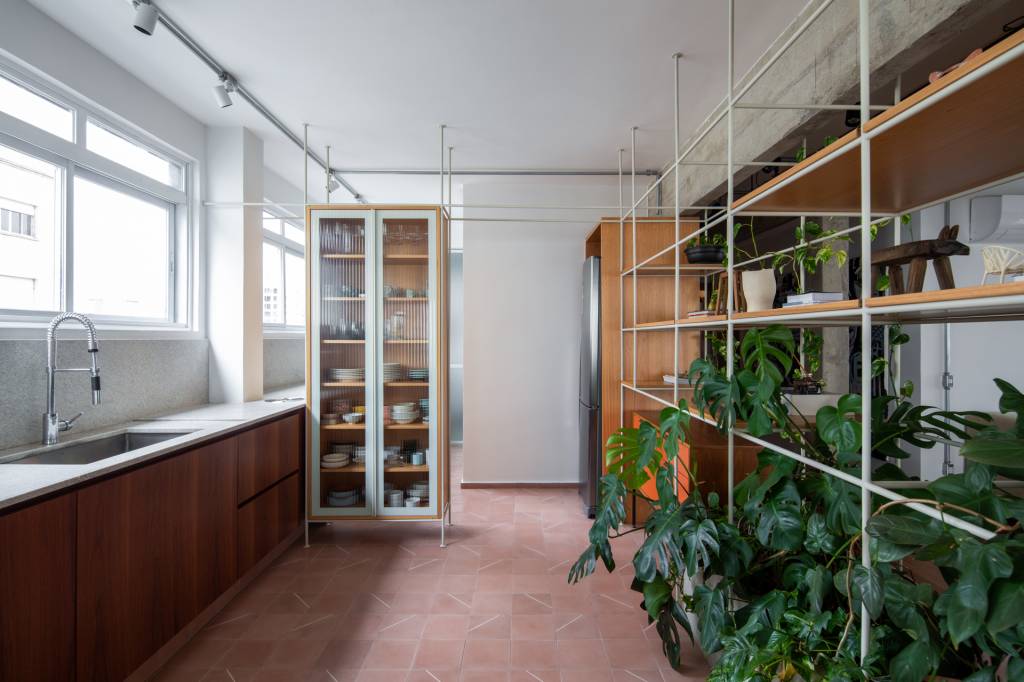 Neste apê, estante metálica branca é divisória e floreira para jiboias e costelas-de-adão. Projeto de Península Arquitetura.