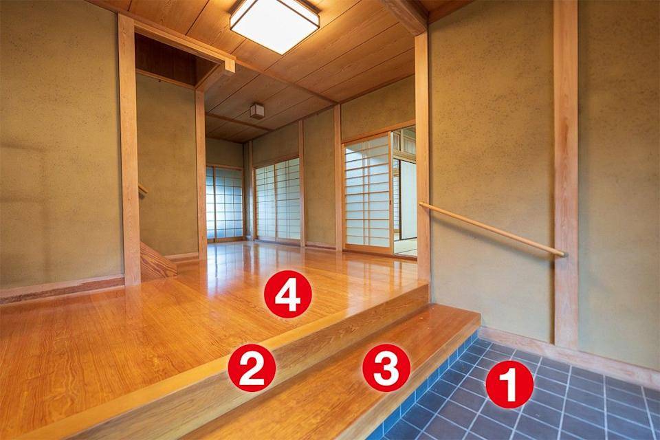 Entrada de uma casa japonesa com piso de cer&acirc;mica escura, dois degraus de madeira clara e um corrim&atilde;o simples. Ao fundo, piso de madeira brilhante e portas de correr shoji com treli&ccedil;as de madeira e papel transl&uacute;cido, iluminadas por luz natural e lumin&aacute;rias retangulares no teto de madeira