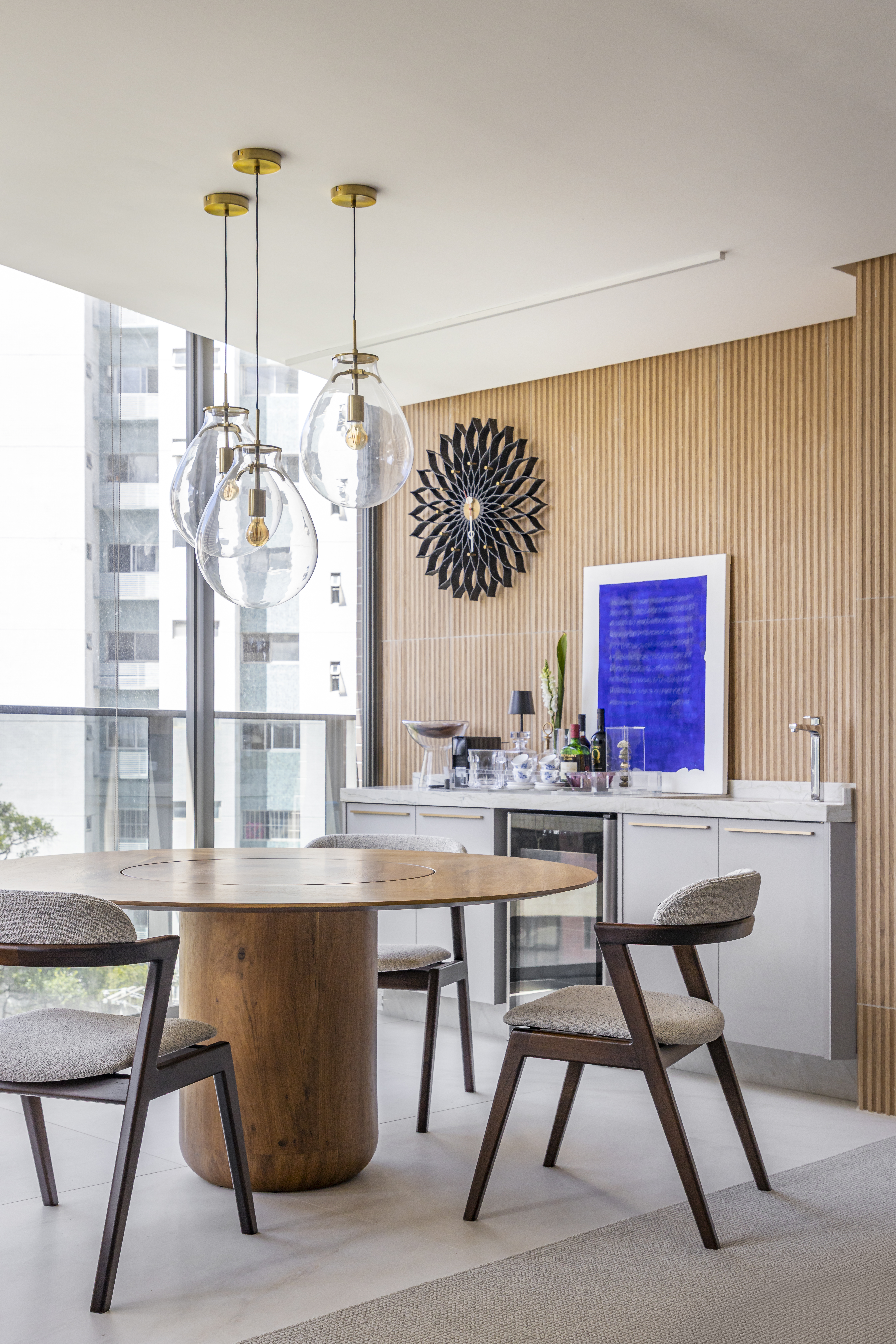 Apartamento em Recife &eacute; transformado em ref&uacute;gio urbano sofisticado e atemporal. Projeto de Rom&aacute;rio Rodrigues Arquitetura. Na foto, Sala de jantar moderna com mesa redonda de madeira clara e quatro cadeiras estofadas cinzas. Acima da mesa, tr&ecirc;s pendentes de vidro transparente com detalhes dourados. Ao fundo, parede ripada de madeira, balc&atilde;o cinza com pia, adega e bebidas, e um quadro azul vibrante. Uma janela grande revela pr&eacute;dios ao longe